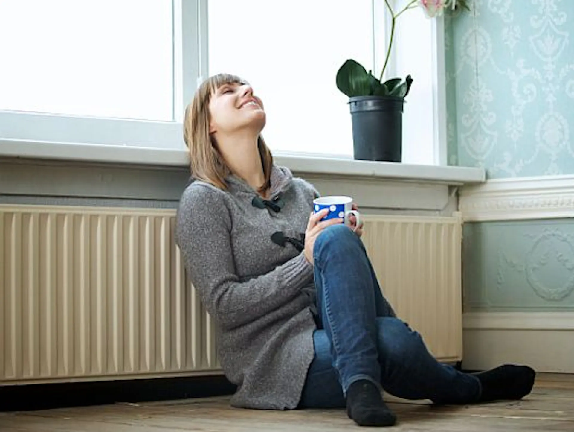 Frau sitzt lächelnd mit einer Tasse vor dem Heizkörper unter dem Fenster