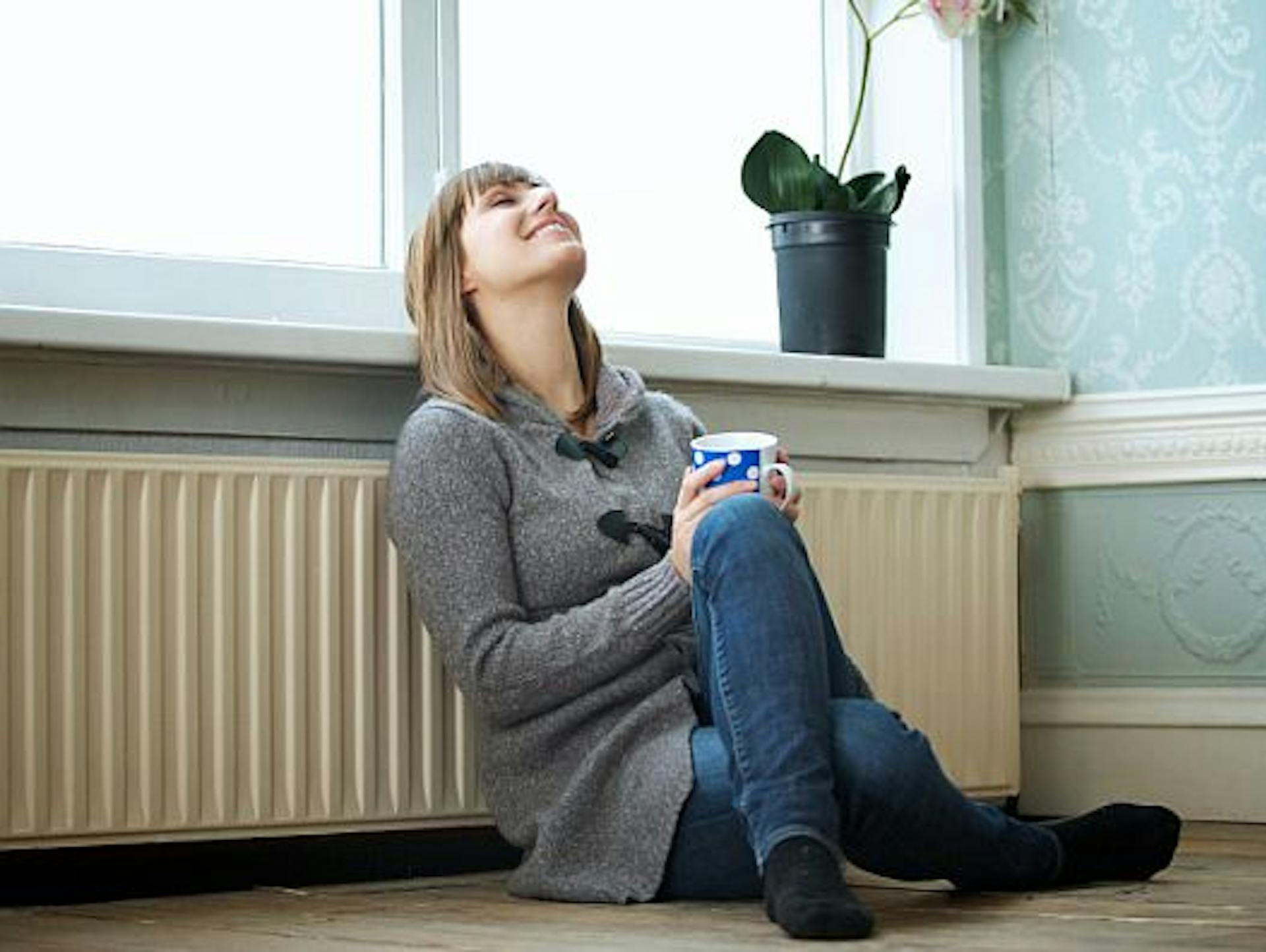 Donna sorridente con una tazza seduta di fronte al radiatore sotto la finestra