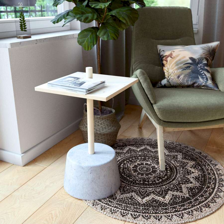 Ein Beistelltisch mit einem Betonsockel steht neben einem dunkelgrünen Sessel vor einer Pflanze auf einem kleinen, runden Teppich