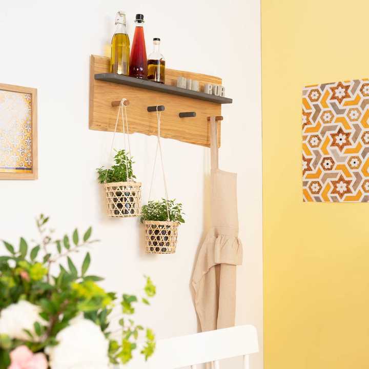 An einer weißen Wand hängt Garderobe Gerda, an ihr hängen Pflanzen sowie eine Schürze. Obendrauf stehen Flaschen.