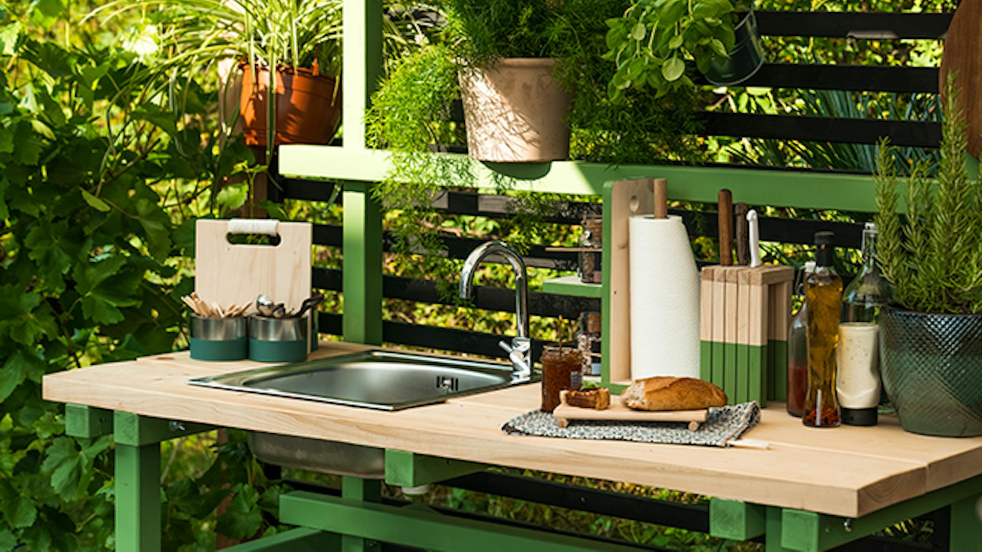 Outdoorküche Alfons, welche hellgrün gestrichen worden ist, steht in einer Ecke eines sommerlichen Gartens.