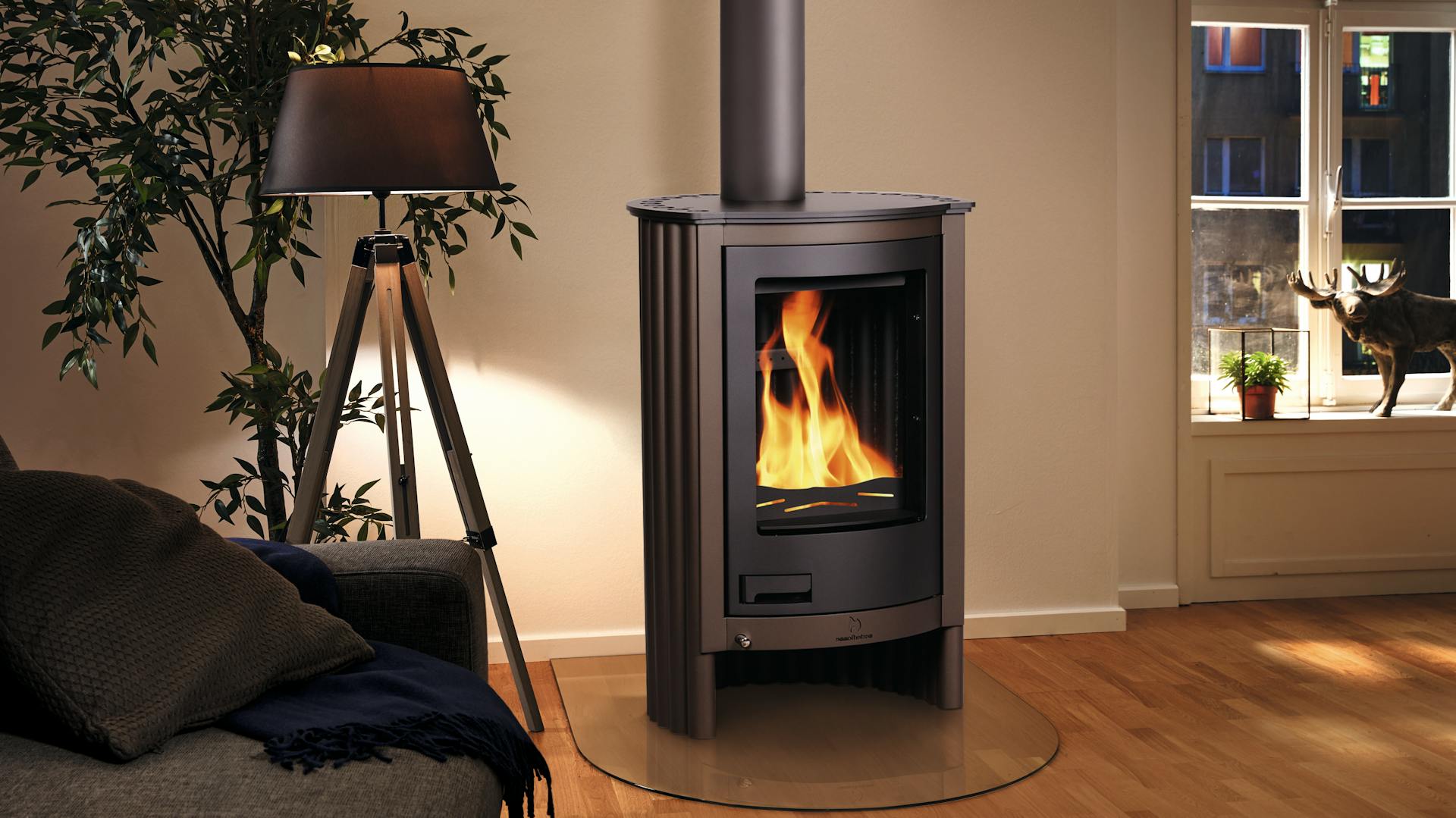 EIn moderner Holzofenkamin steht in einem Wohzimmer mit Holzboden neben einer Stehtlampe und einem Fenster.