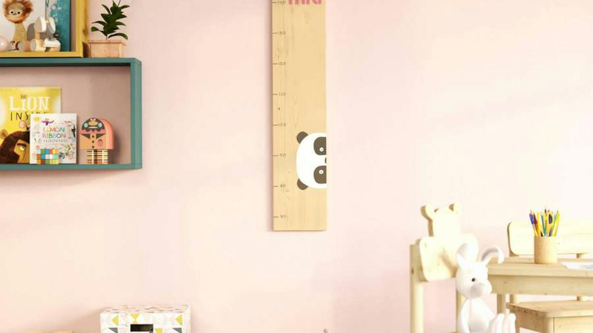 Eine Kindermesslatte mit Pandamotiv hängt an einer rosa farbigen Wand. Neben und vor der Messlatte befinden sich weitere Kindermöbel.