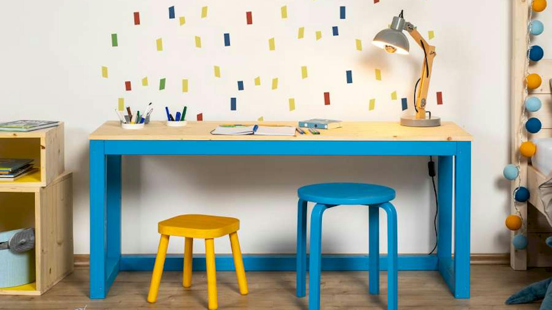 Ein bunter Schreibtisch und bunte Stühle aus Holz stehen vor einer Wand mit Tape-Art.