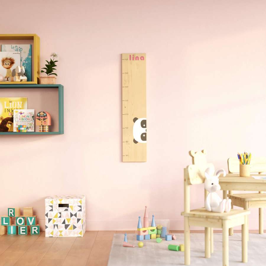 Eine Kindermesslatte mit Pandamotiv hängt an einer rosa farbigen Wand. Neben und vor der Messlatte befinden sich weitere Kindermöbel.