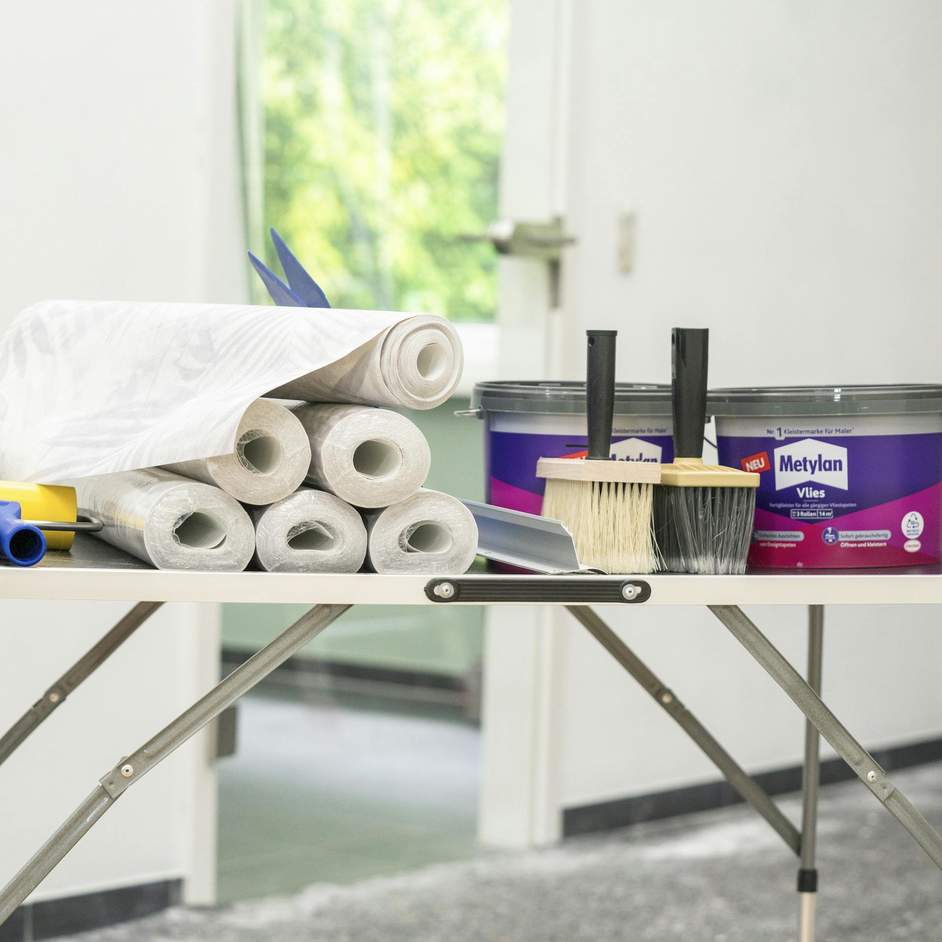 Ein Tapeziertisch, darauf befinden sich Tapetenrollen, Kleber und Malerwerkzeuge.