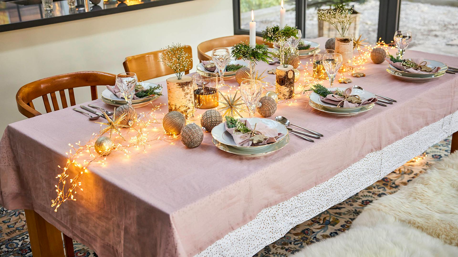 Ein großer Festtagstisch, auf dem Lichterketten, dekorative Weihnachtskugeln und Kerzen in vergoldeten Haltern festlich platziert sind.