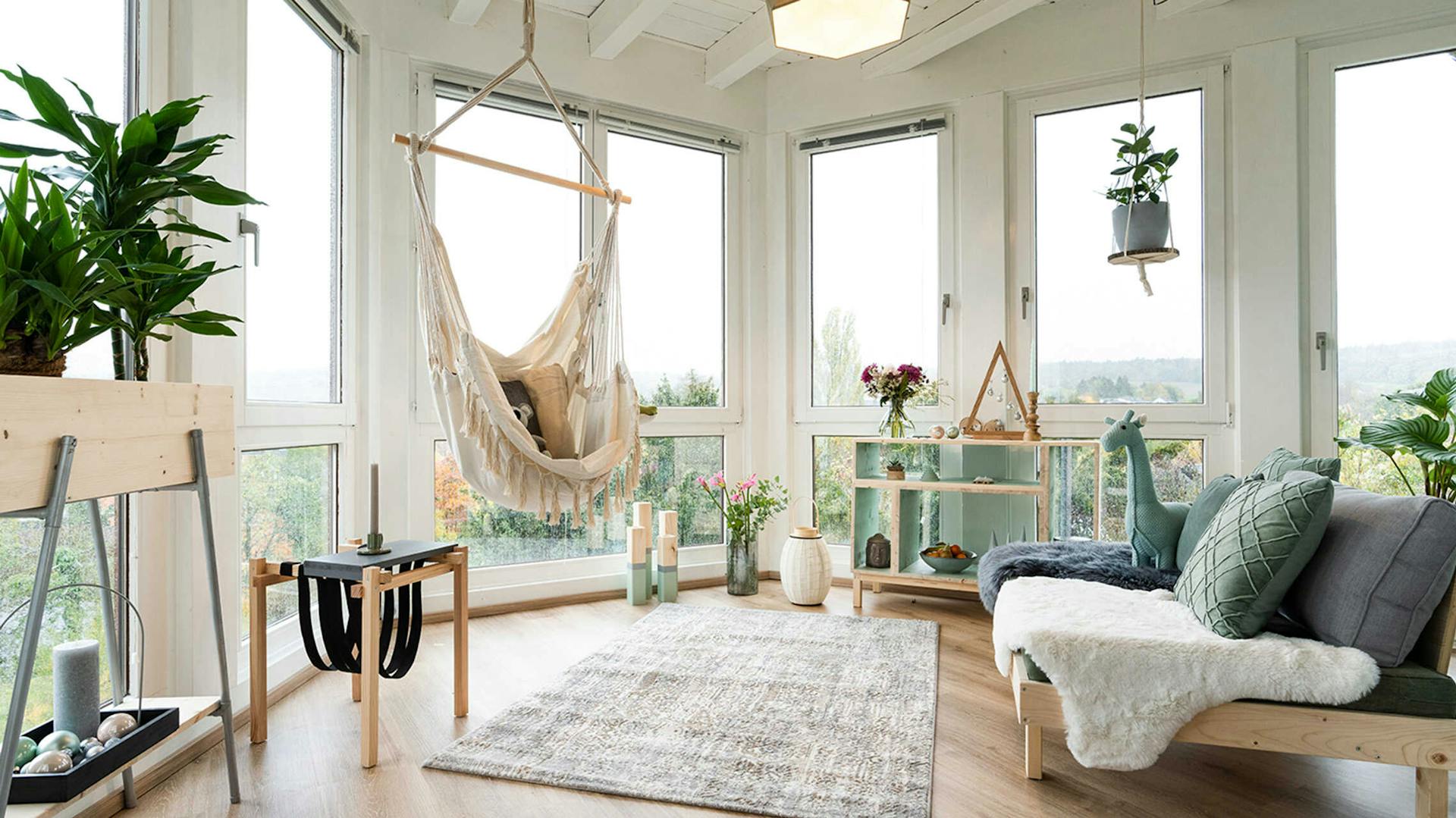 Eine offene Chill-Out-Area wurde in einer Ecke mit vielen Fenstern eingerichtet. Gegenüber von einem Hängesessel steht eine Couch. Außerdem befinden sich in der Ecke ein Regal, ein Beistelltisch und verschiedene Deko-Elemente.