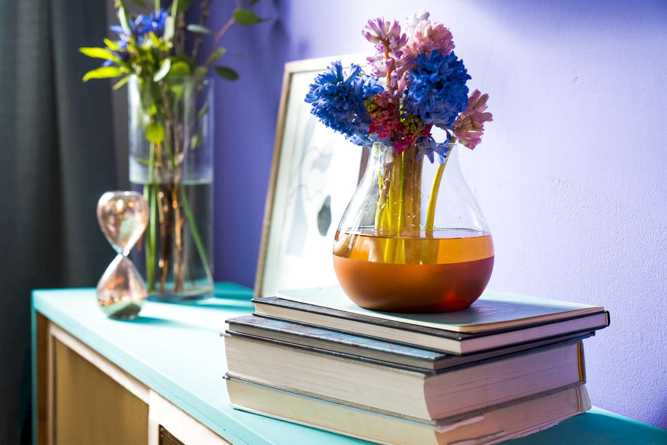 Eine Vase mit Kupferdetail ist Bücher platziert, die auf einem blauen Regal liegen. Im Hintergrund befinden sich eine weitere Vase, ein Bild und eine Sanduhr.
