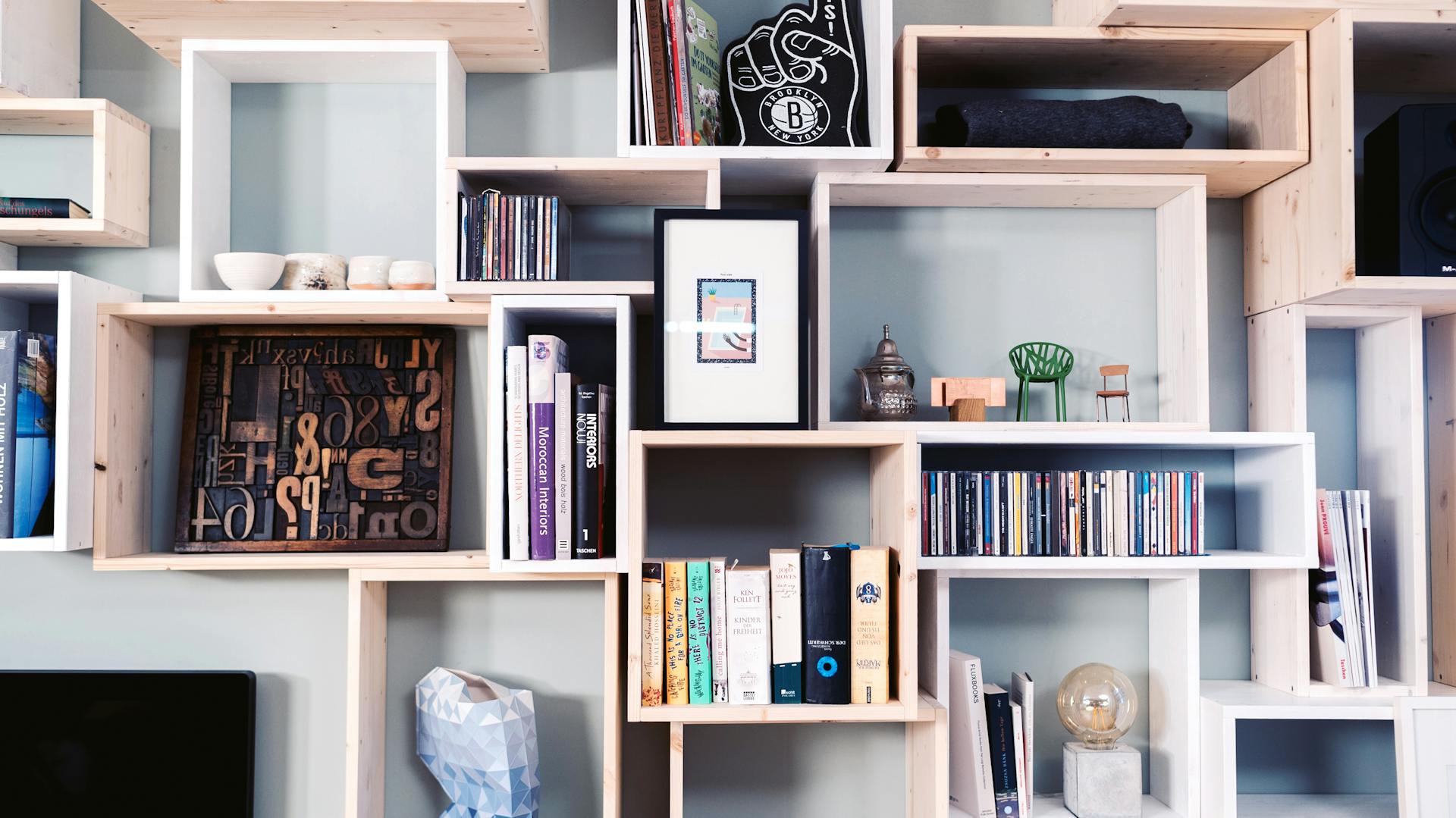 Holzregal mit bunten Flächen mit Dekoartikeln und Büchern sowie ein Bett im Vorder- und ein Schreibtisch im Hintergrund.