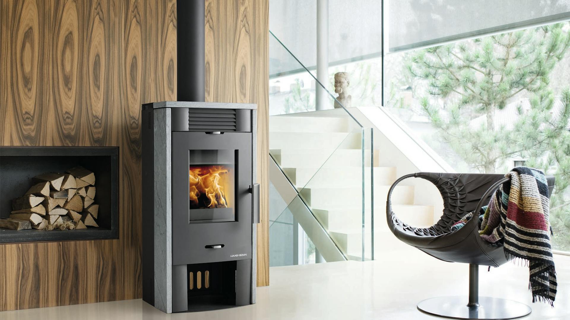 Ein Holzkaminofen mit Holzregal in der Wand brennt in einem modernen, verglasten Wohnzimmer. Rechts steht ein Stuhl mit einer bunten Decke.