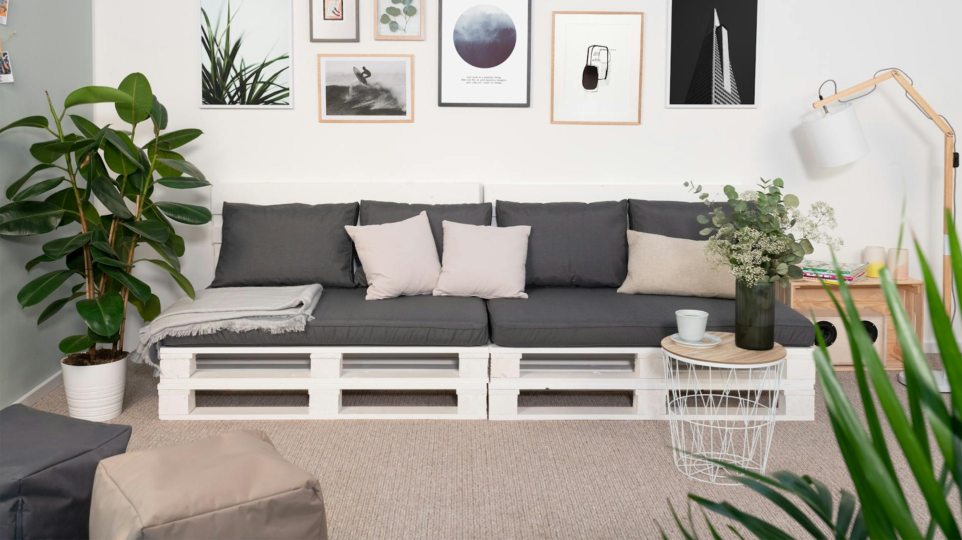 Divano in pallet bianco con cuscini grigi in soggiorno su tappeto davanti a una parete con immagini incorniciate