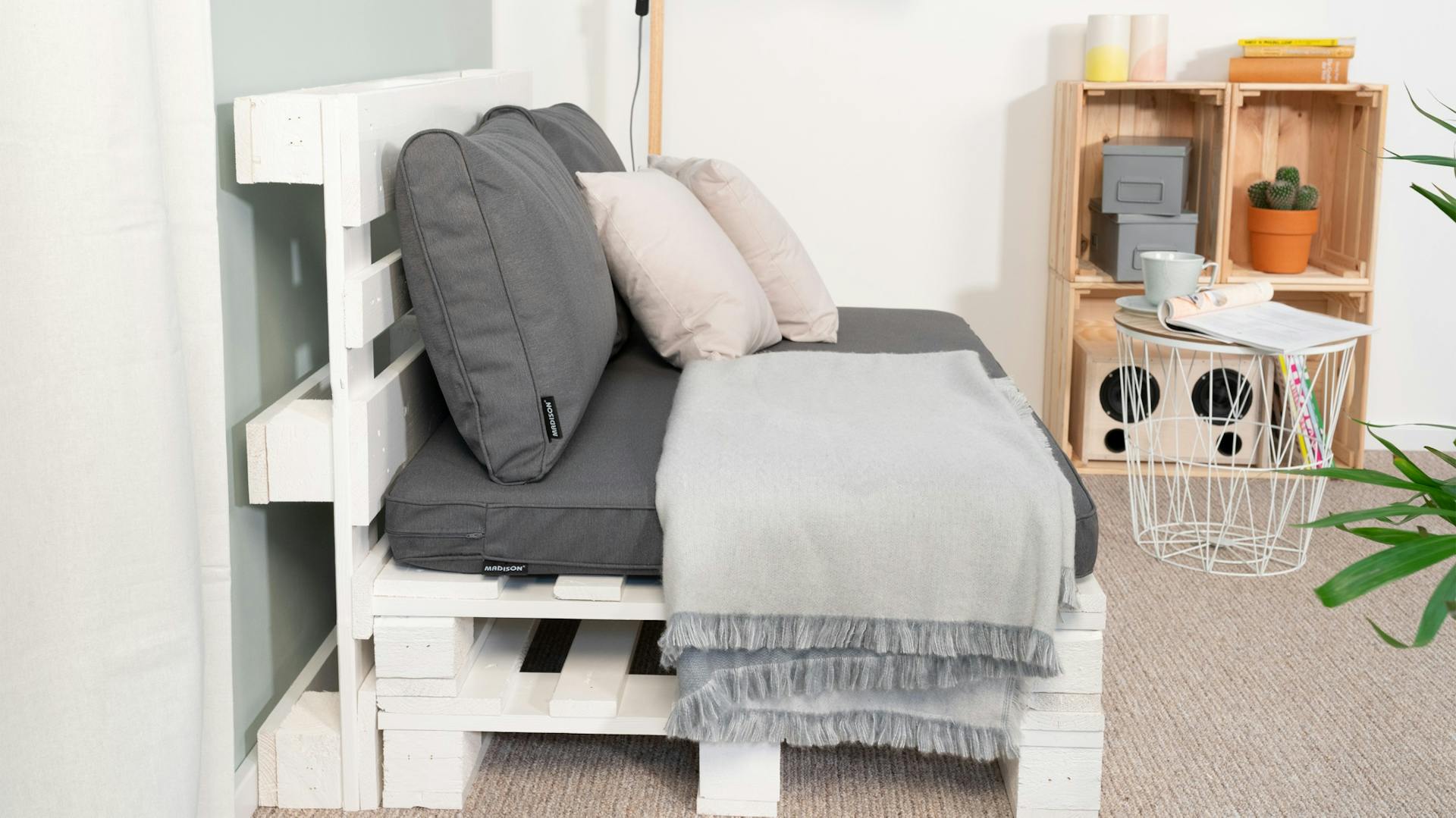 Seitlich fotografiertes Palettensofa mit Auflage, Decke und Kissen auf Teppich vor hellblauer Wand