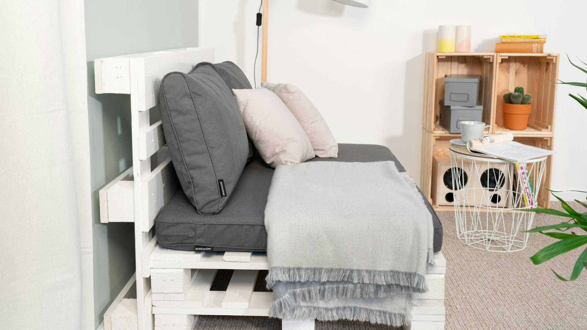 Divano di pallet fotografato lateralmente con rivestimento, coperta e cuscino su tappeto davanti a una parete celeste
