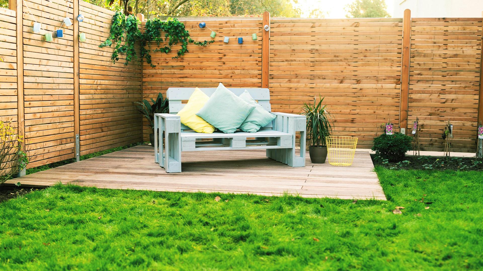 Hellblaues Palettensofa mit Kissen auf Sitzplatz aus Holzdielen in Garten vor Zaun