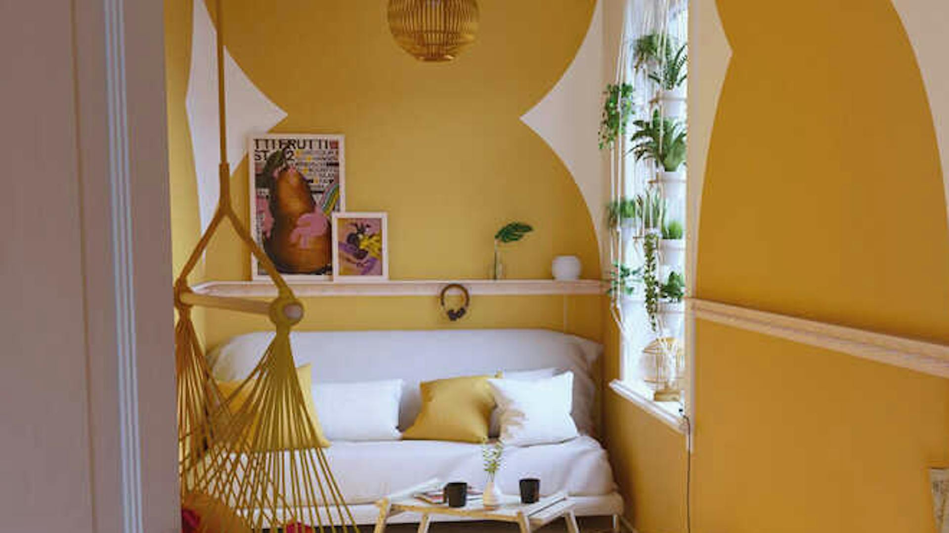 In einem gelb gestalteten Raum steht an der hinteren Wand eine helle Couch mit weißen und gelben Kissen. Davor sind Couchtisch Philip und ein weißer Pouf platziert und ein Hängesessel angebracht.