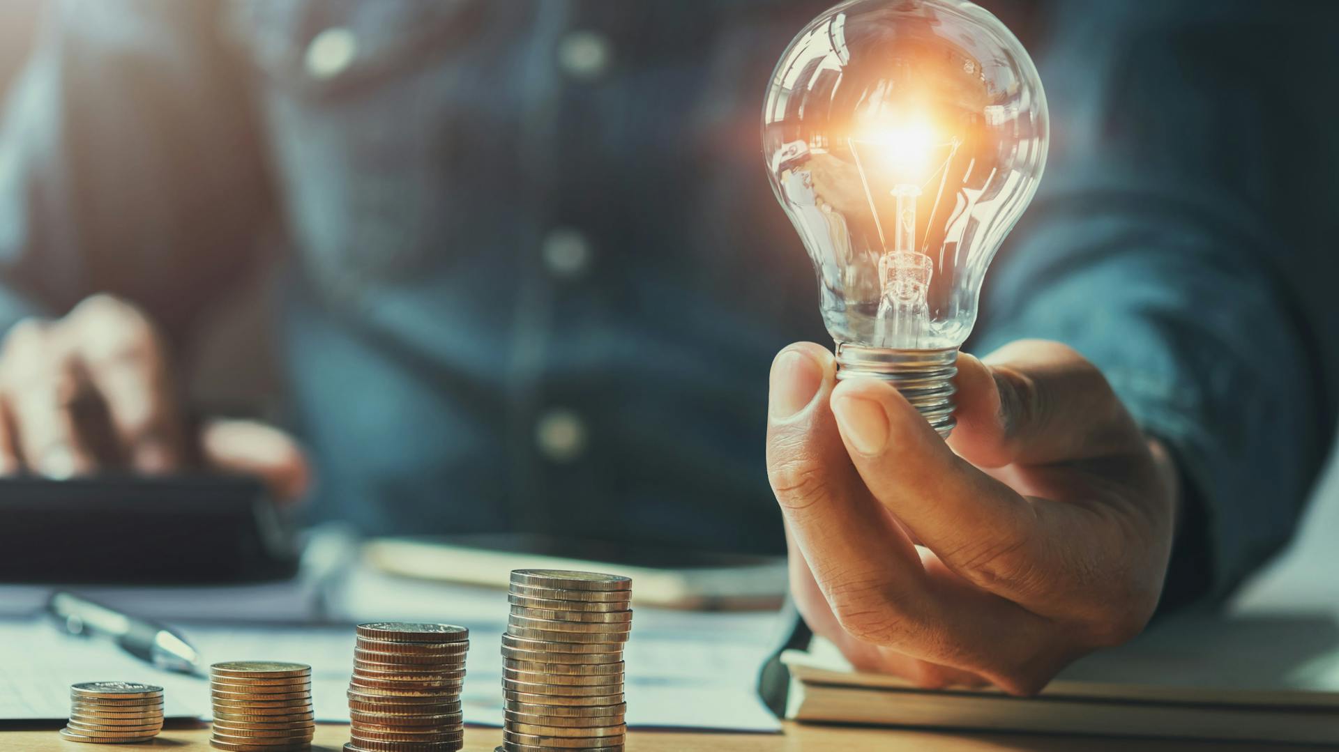 Person hält leuchtende Glühbirne in der Hand neben Münzen, die auf einem Tisch zu Türmen gestapelt sind.