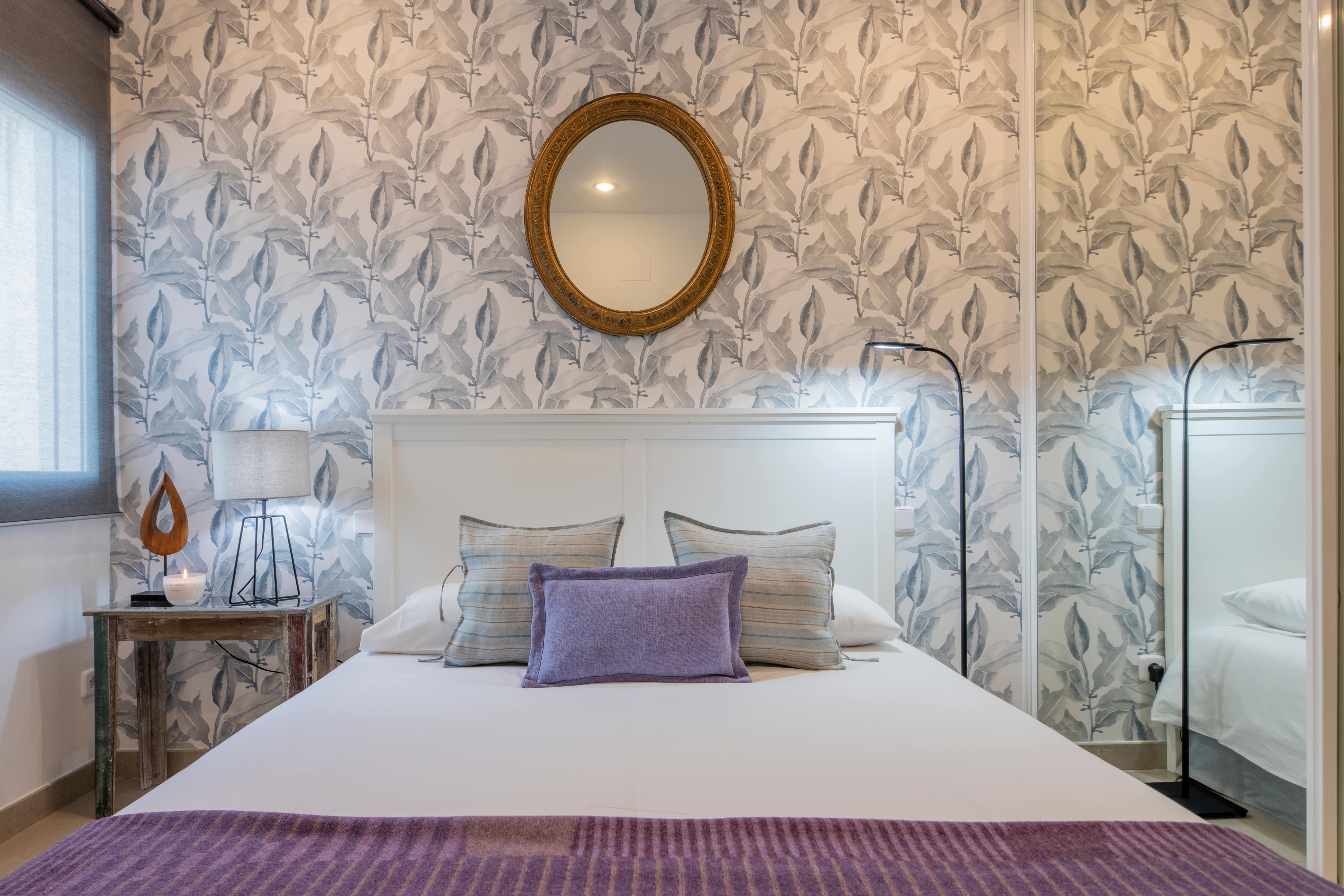 Bett vor einer Wand mit floraler Tapete. Daran hängt ein brauner Spiegel.