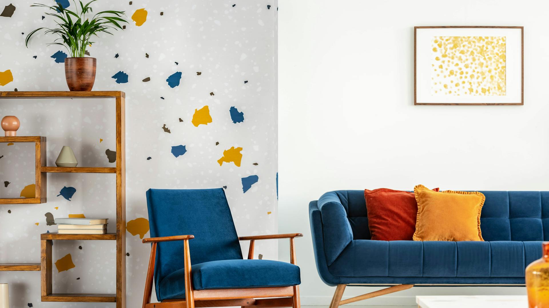 Tapete mit auffälligem Muster in blau-gelb an Wohnzimmerwand mit Sofa, Pflanze und Bücherregal.