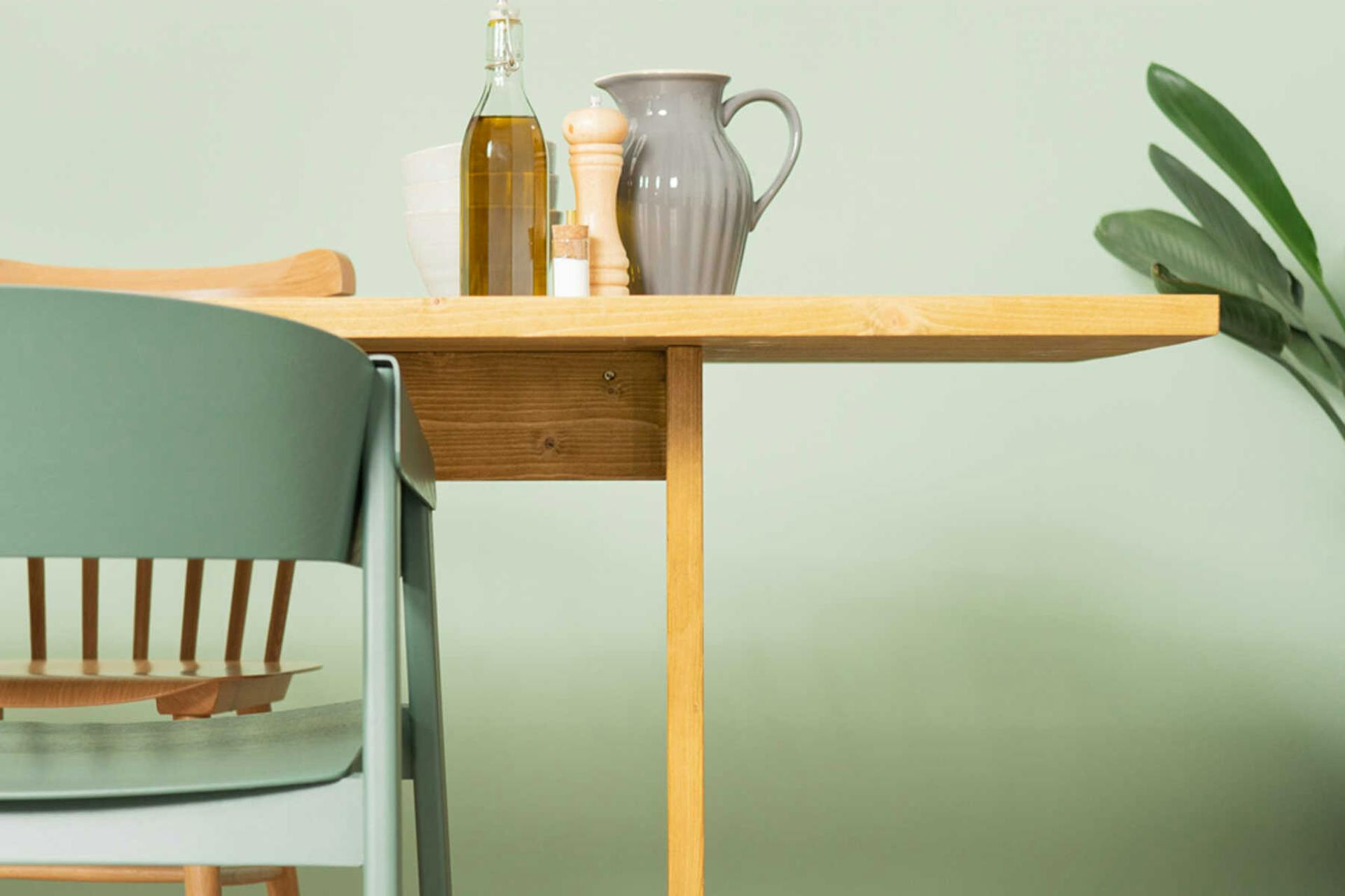 Tisch August steht vor einer pastellgrünen Wand. Auf dem Tisch steht Geschirr und Küchenutensilien.