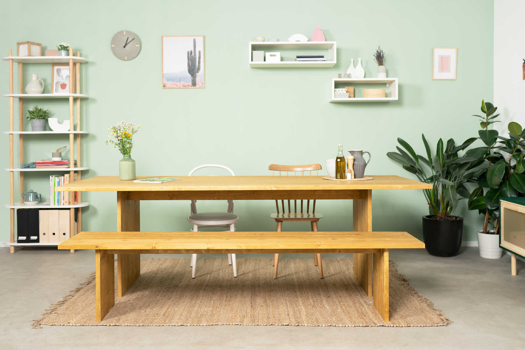 Tisch August steht in einem Raum mit einer pastellgrünen Wand. Vor dem Tisch steht eine passende Bank.