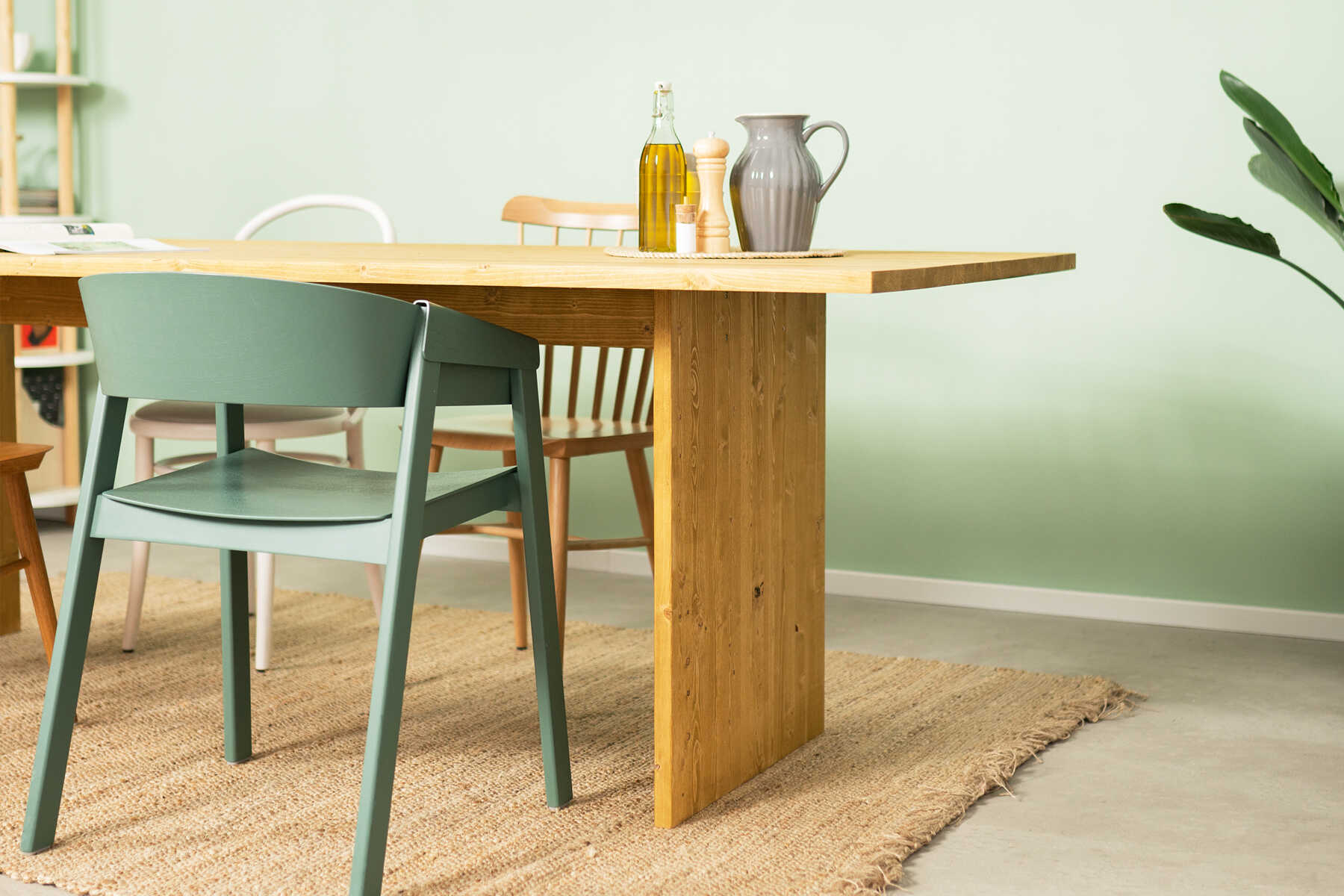 Tisch August steht, umgeben von Stühlen, vor einer pastellgrünen Wand.