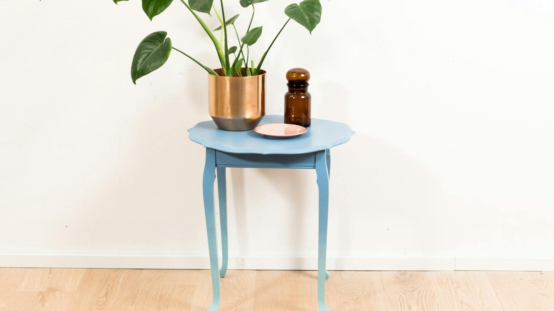 Odkládací stolek s barevným přechodem od modré k světle modré. Na něm stojí dekorace a pokojová rostlina.