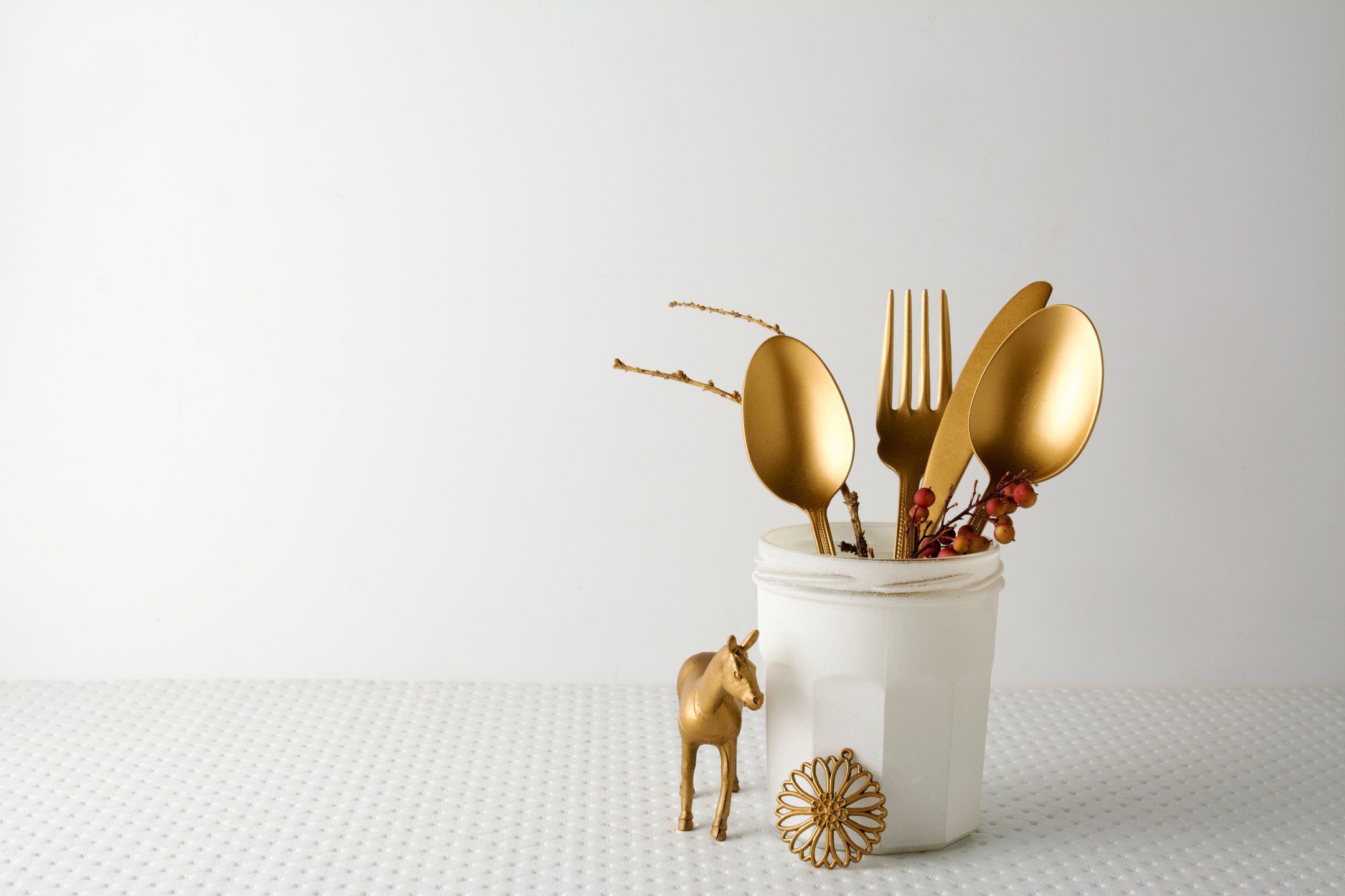 Un vasetto di marmellata dipinto di bianco funge da portaposate per posate dorate.