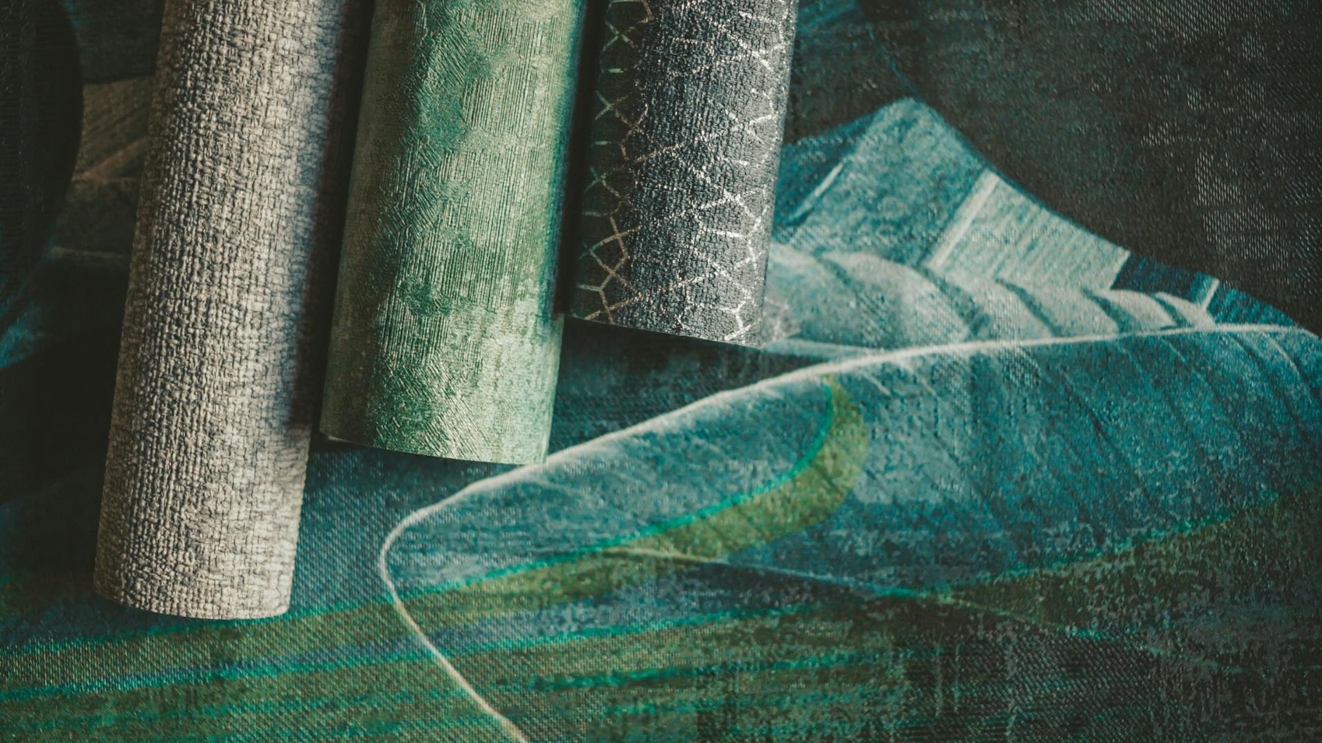 Drei verschiedene strukturierte Tapetenrollen in Grüntönen liegen auf einer ausgerollten Tapetenbahn mit Blattmuster.