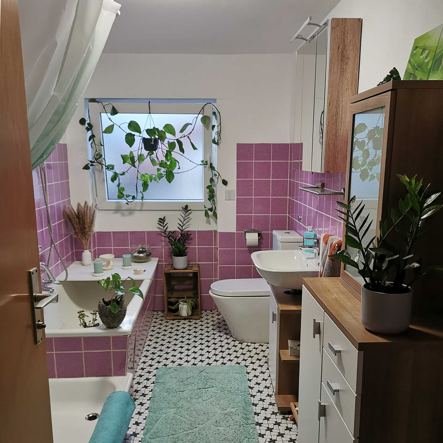 In einem Badezimmer mit pinken Fliesen befindet sich eine Badewanne unter dem Fenster, vor der eine Duschwanne platziert ist. Auf der gegenüberliegenden Seite befinden sich eine Toilette, ein Waschbecken und einige Schränke.