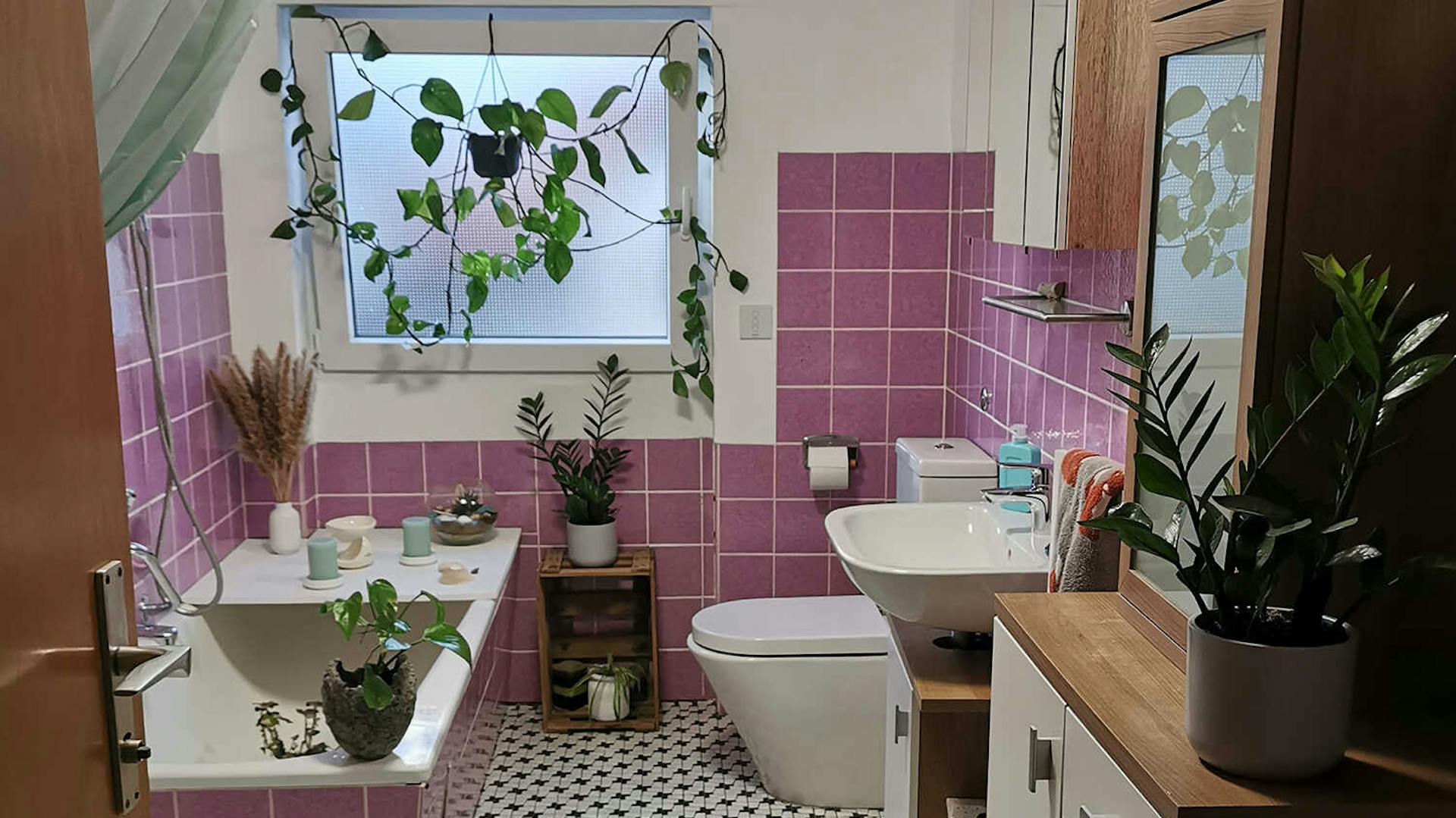 In einem Badezimmer mit pinken Fliesen befindet sich eine Badewanne unter dem Fenster, vor der eine Duschwanne platziert ist. Auf der gegenüberliegenden Seite befinden sich eine Toilette, ein Waschbecken und einige Schränke.