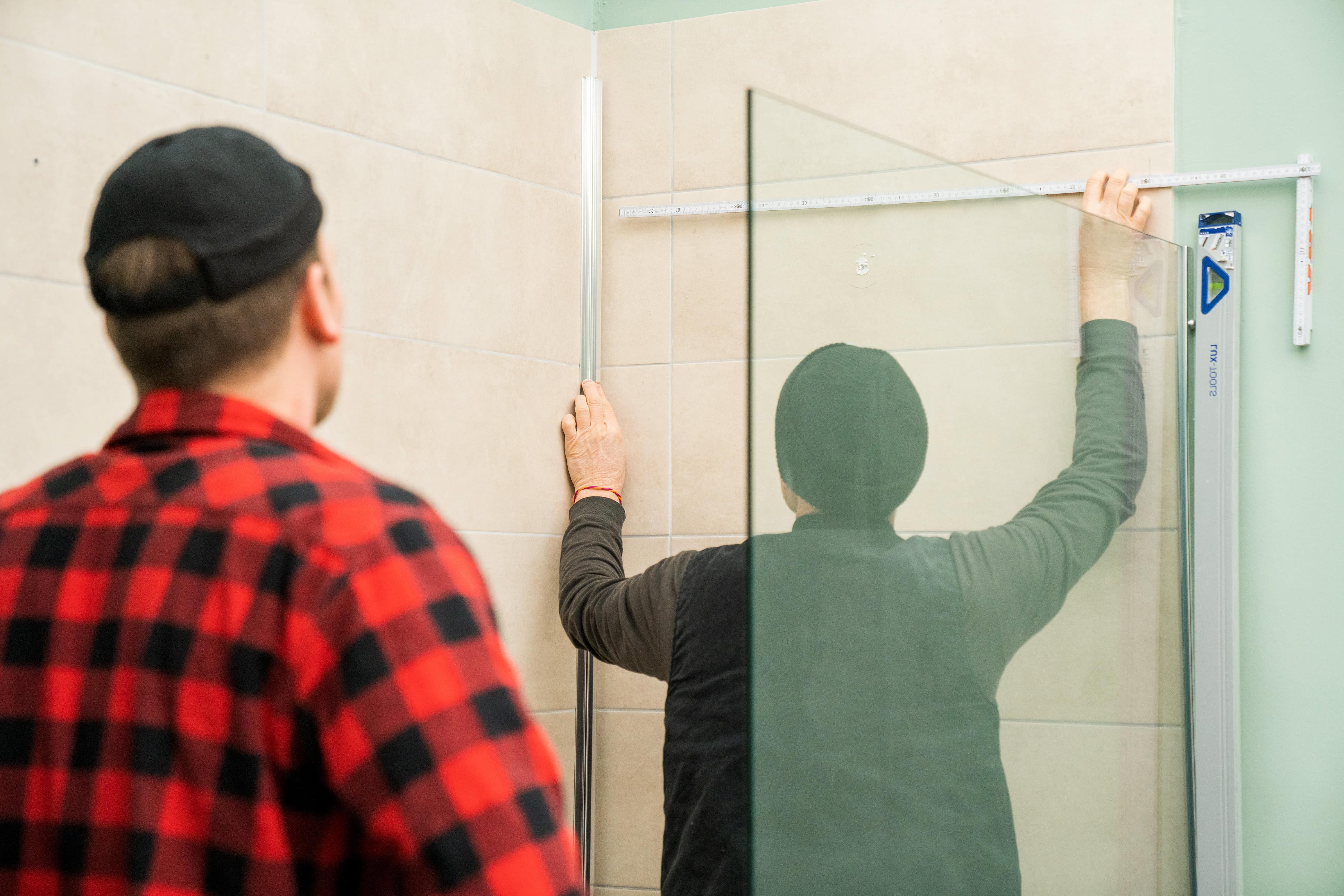 Zwei Handwerker messen die Breite einer Dusche mit Fliesenspiegel aus.
