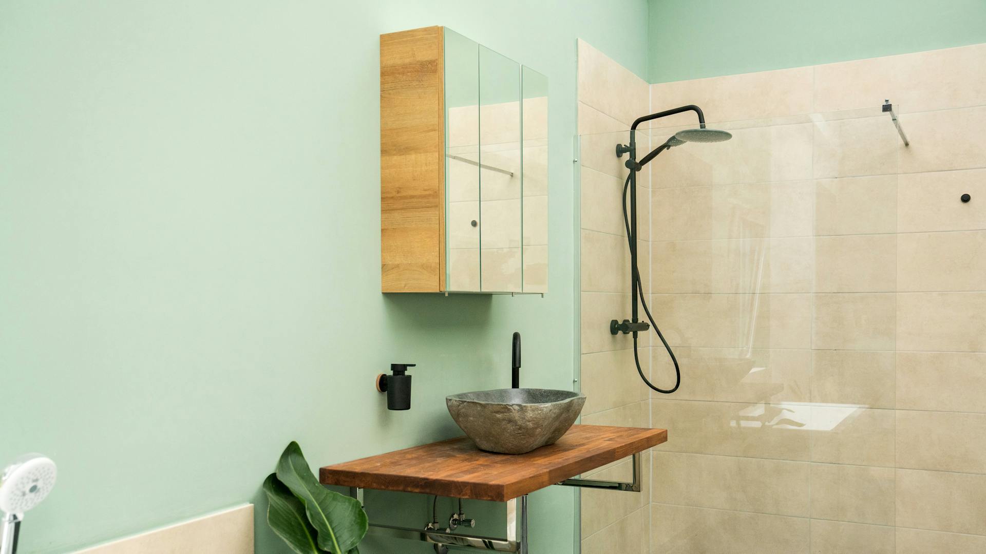 Spiegelschrank hängt über einem Waschtisch mit holzplatte und grauem Waschbecken in einem Badezimmer mit hellgrünen Wänden und beige gefliester Dusche.