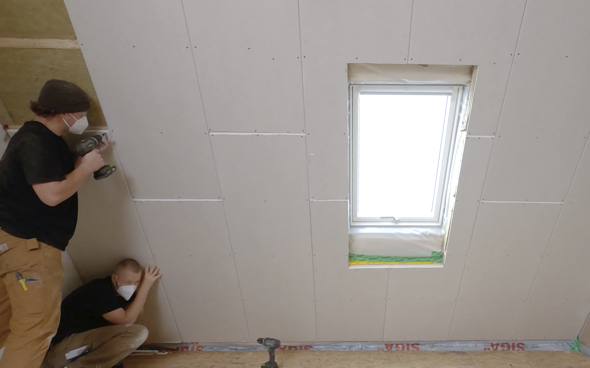 Zwei Handwerker stehen an einer Dachschräge und schrauben Gipskartonplatten an. 
