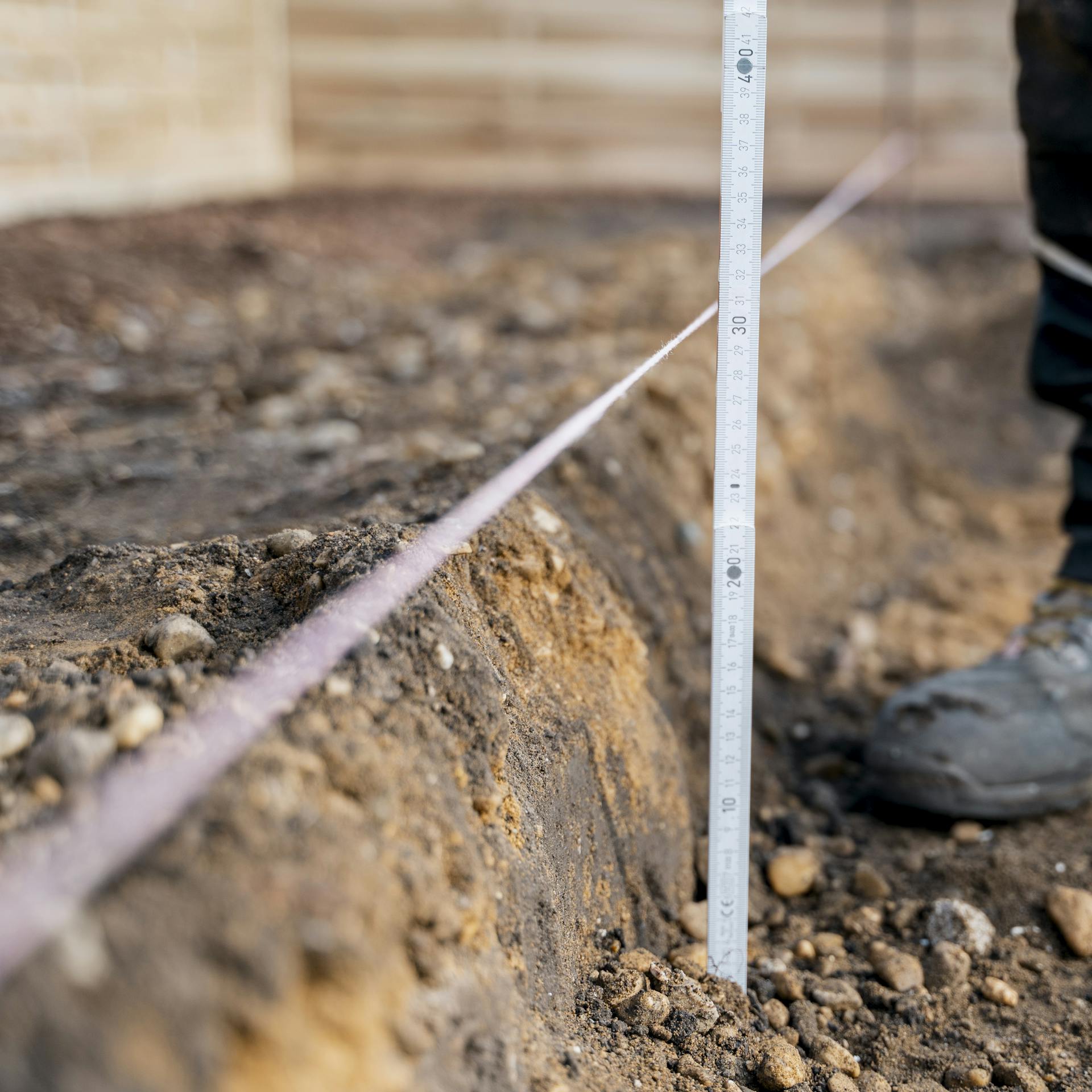 Heimwerker setzt Meterstab an ausgehobenen Boden und Schnurgerüst an.