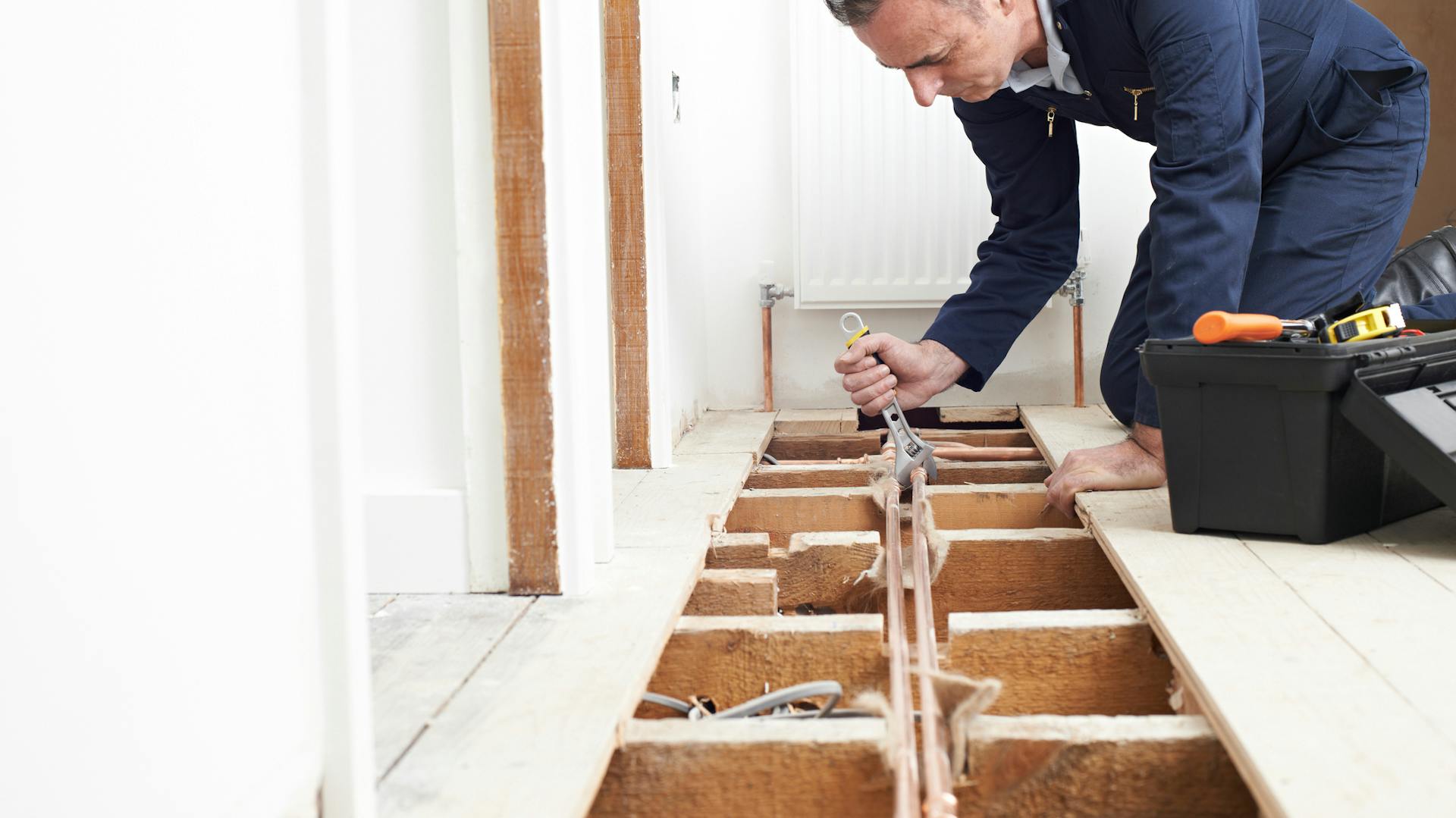 Ein Handwerker kniet an geöffnetem Holzboden und bearbeitet ein Rohr mit einem Schraubenschlüssel.