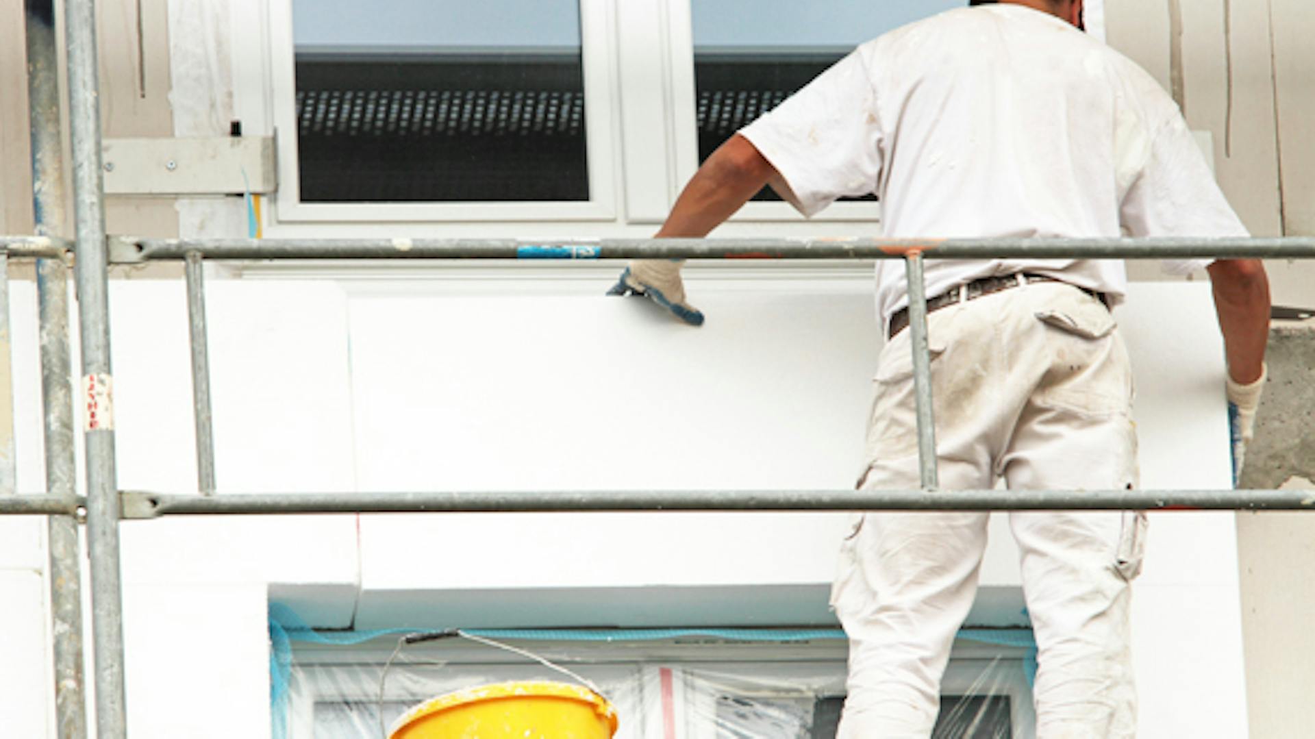 Az ezermester a ház falán állványzaton áll, és szigetelőanyagot tesz a homlokzatra.