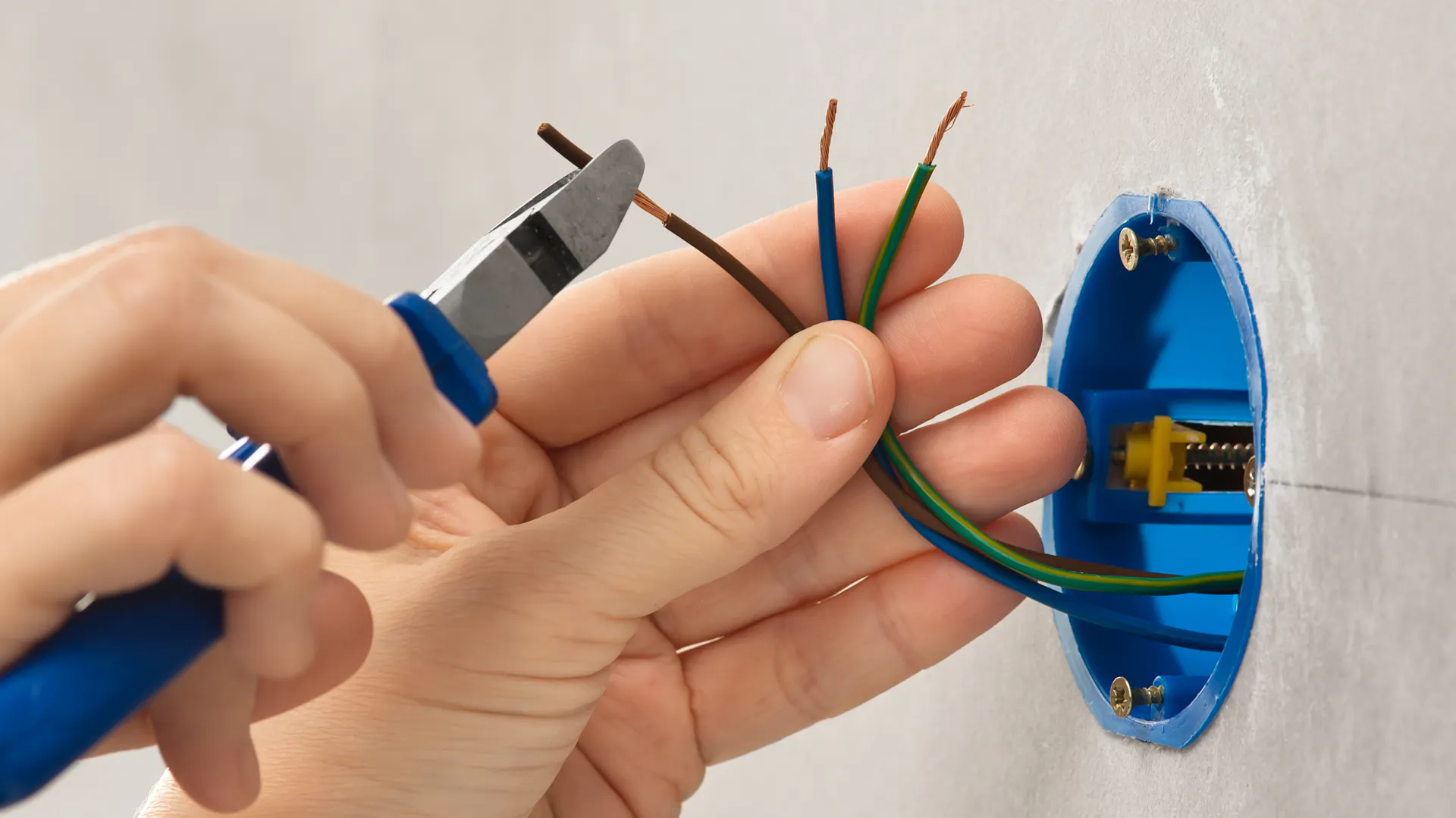 Eine Hand hält eine Zange an Stromkabel, die aus der Wand gucken.