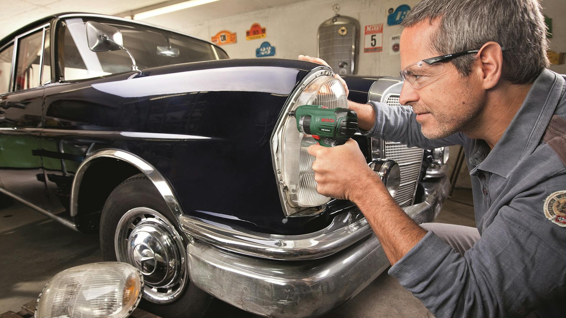 Heimwerker mit Akkuschrauber am Vorderlicht eines Oldtimers.