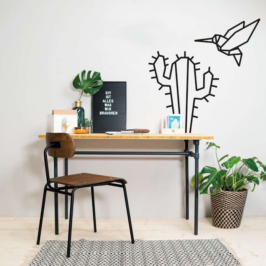 Vor einer weißen Wand mit einen Kaktus und einem Kolibri aus Tape Art steht ein Schreibtisch und ein Stuhl.