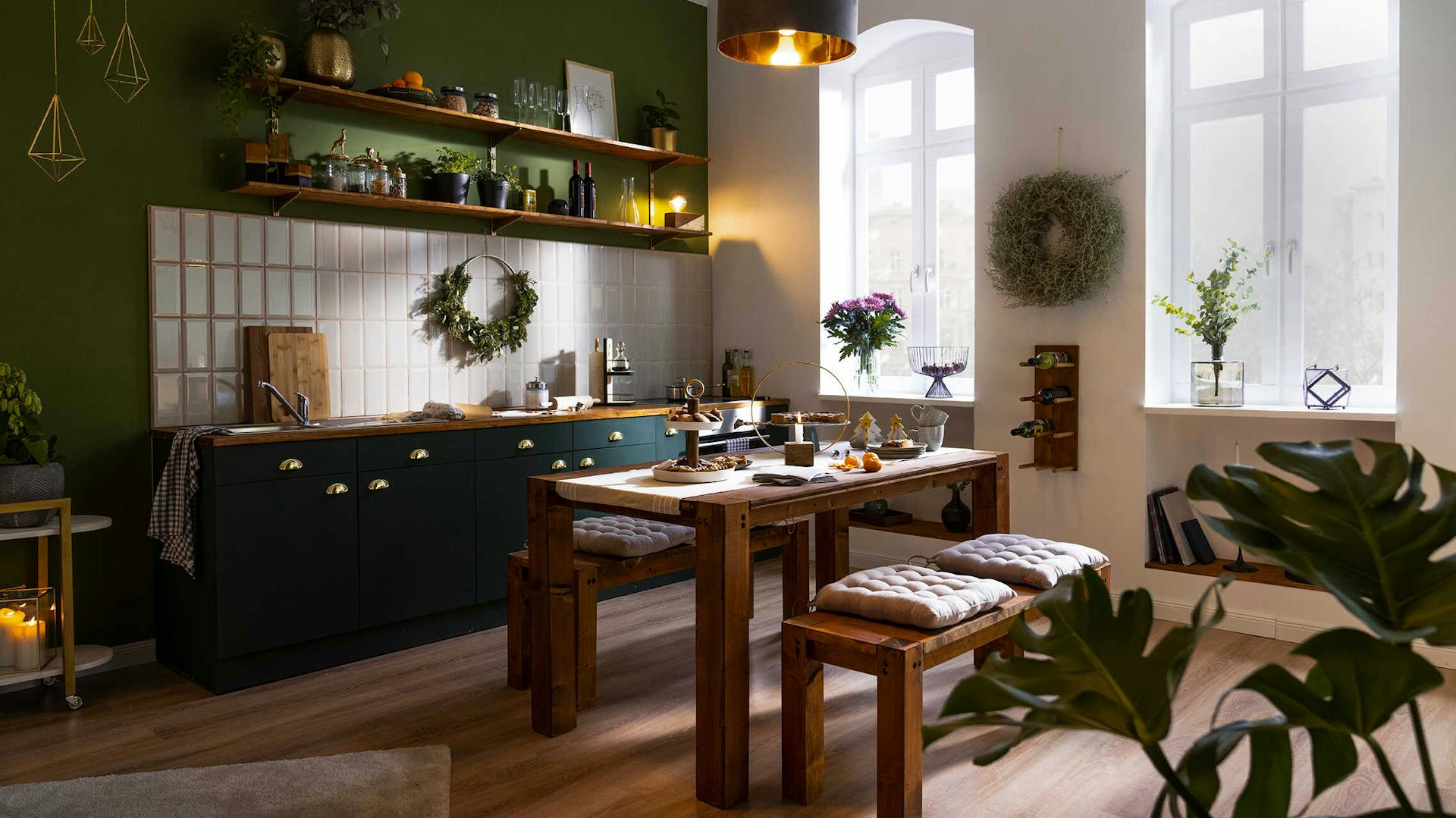 Tisch Maren in dunklem Holz steht mittig in einer großen Küche, die eine einzelne große Wand in Jungle Green besitzt und weiße Metrofliesen als Küchenrückwand nutzt.