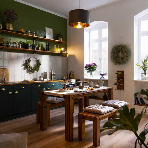 Eine Küche mit grünen Farben. In der Mitte des Raums steht ein großer Holztisch mit Sitzbänken.