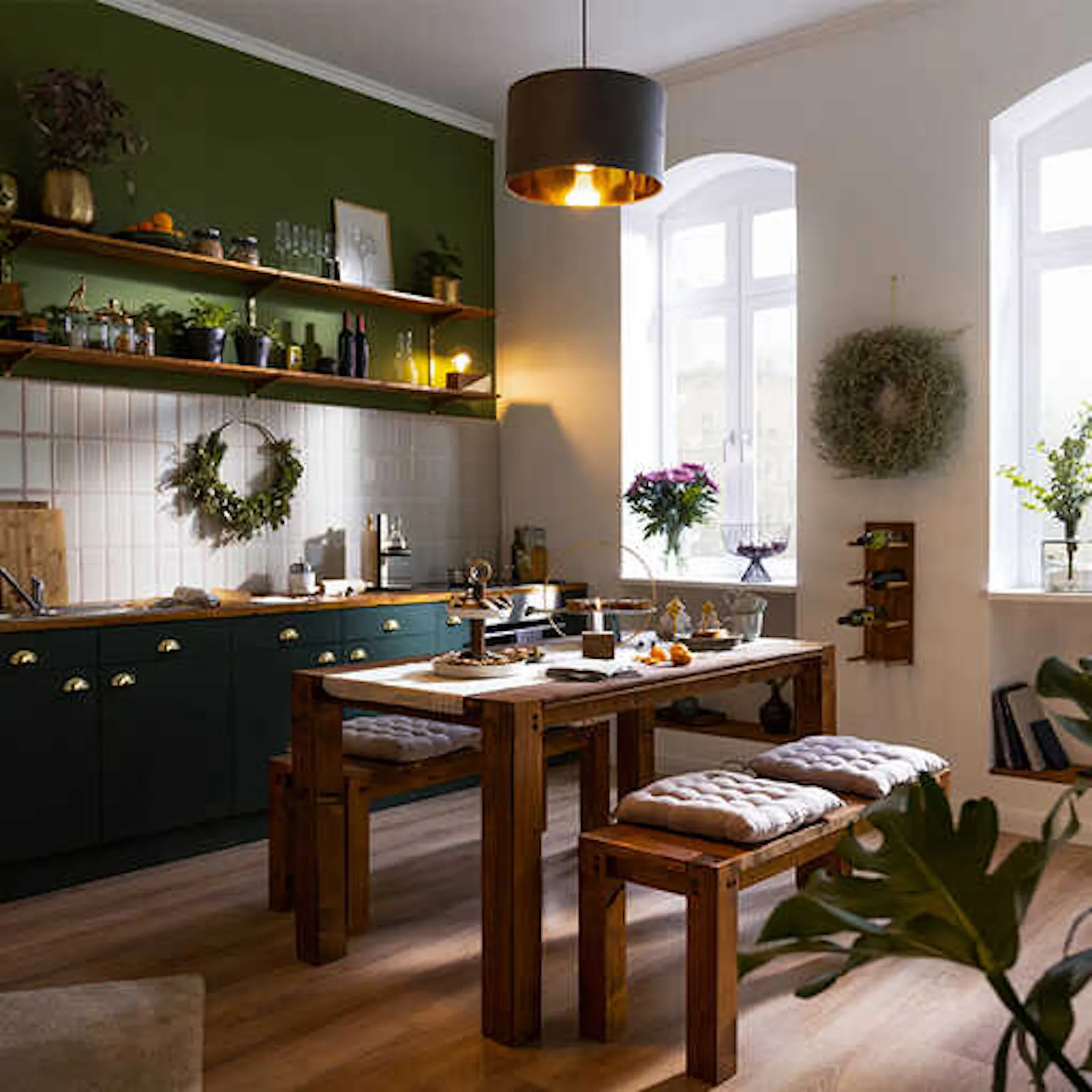 Eine Küche mit grünen Farben. In der Mitte des Raums steht ein großer Holztisch mit Sitzbänken.