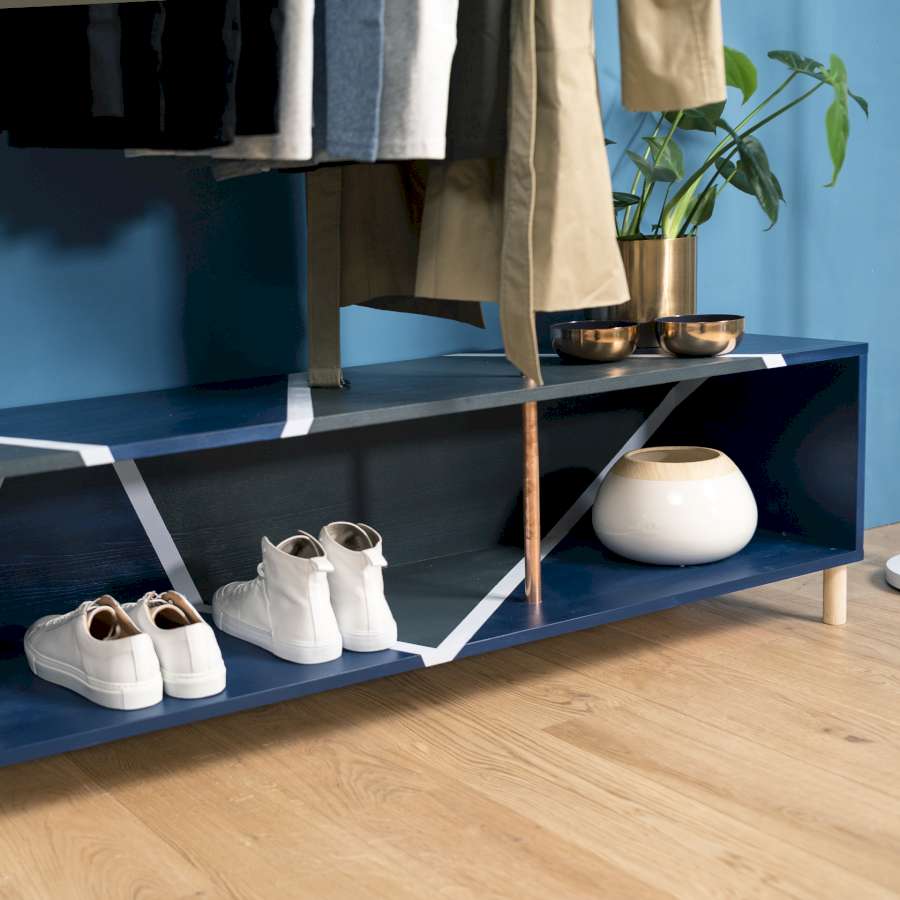 Ein blaues Regal auf dem mit dunkler und weißer Farbe geometrische Muster gemalt wurden. In dem Regal stehen Schuhe und eine Vase und darüber befinden sich eine Pflanze und Jacken.