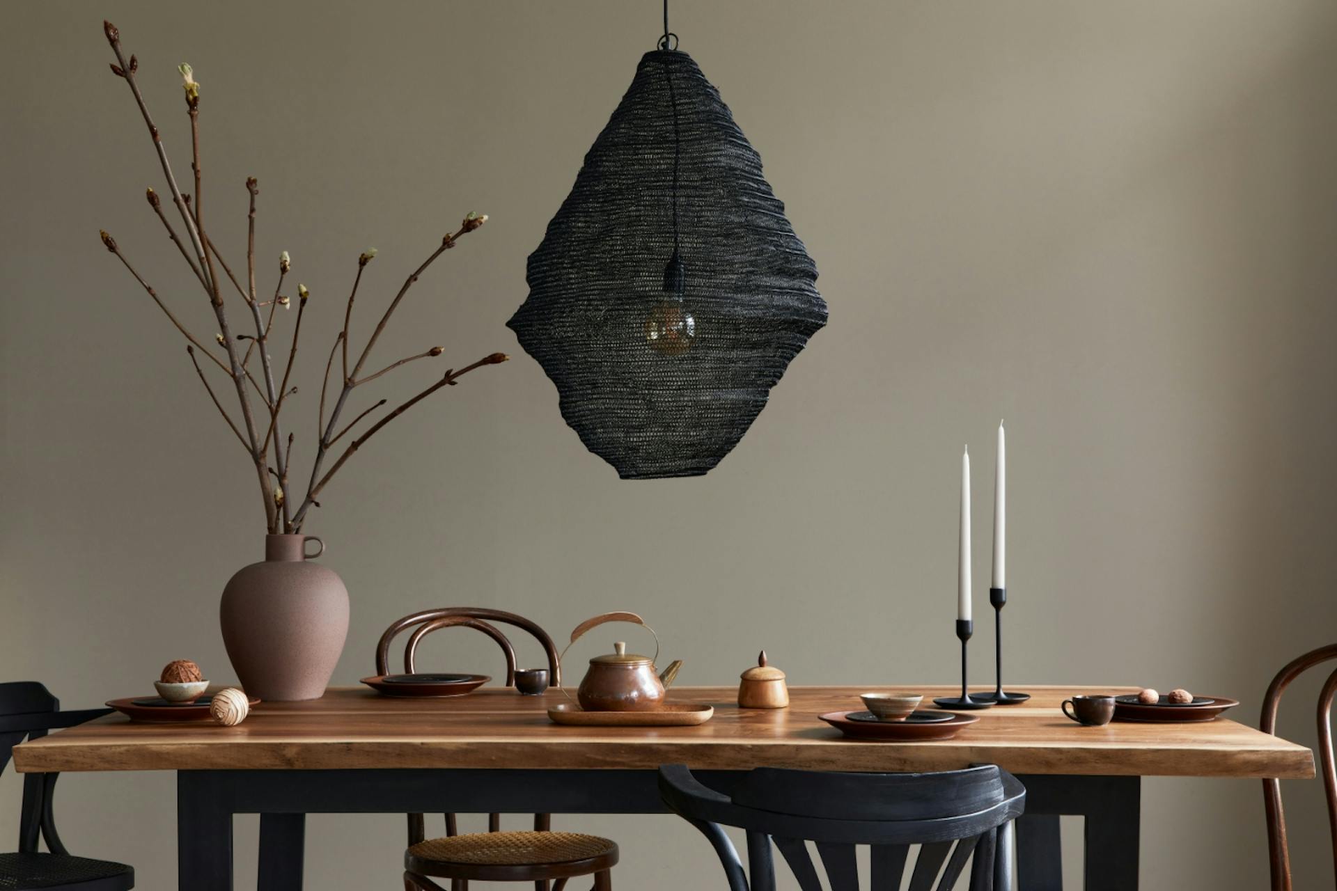Étkezőasztal gyertyákkal, vázával és modern lámpával barnásszürke fal előtt