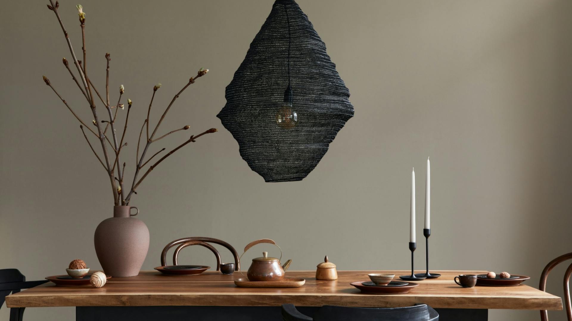 Tavolo da pranzo con candele, vaso e lampada moderna davanti a una parete grigio-marrone