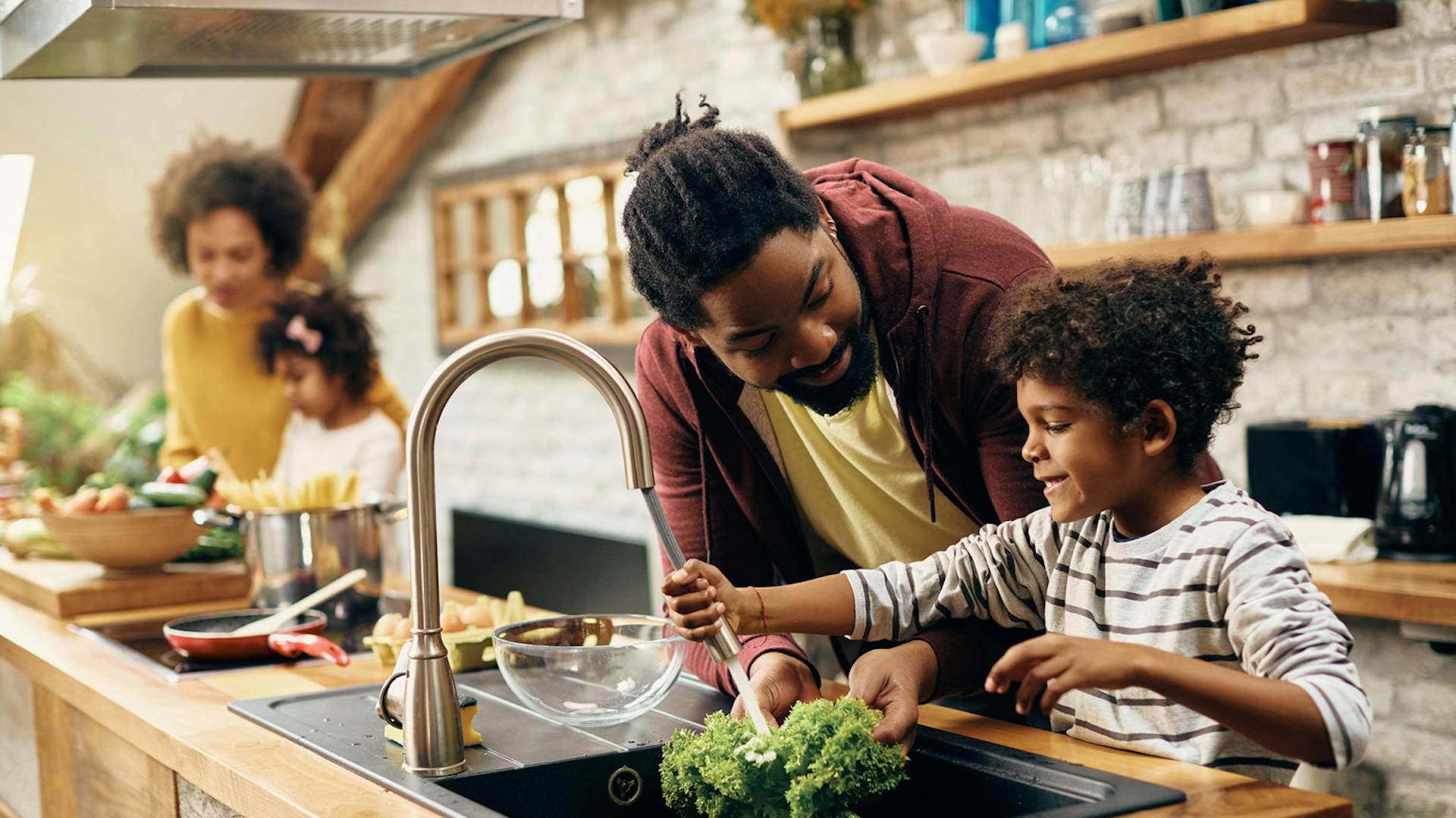 Familie kocht gemeinsam in Küche, im Vordergrund Vater mit Sohn, die Salat abwaschen, und im Hintergrund Mutter mit Tochter.