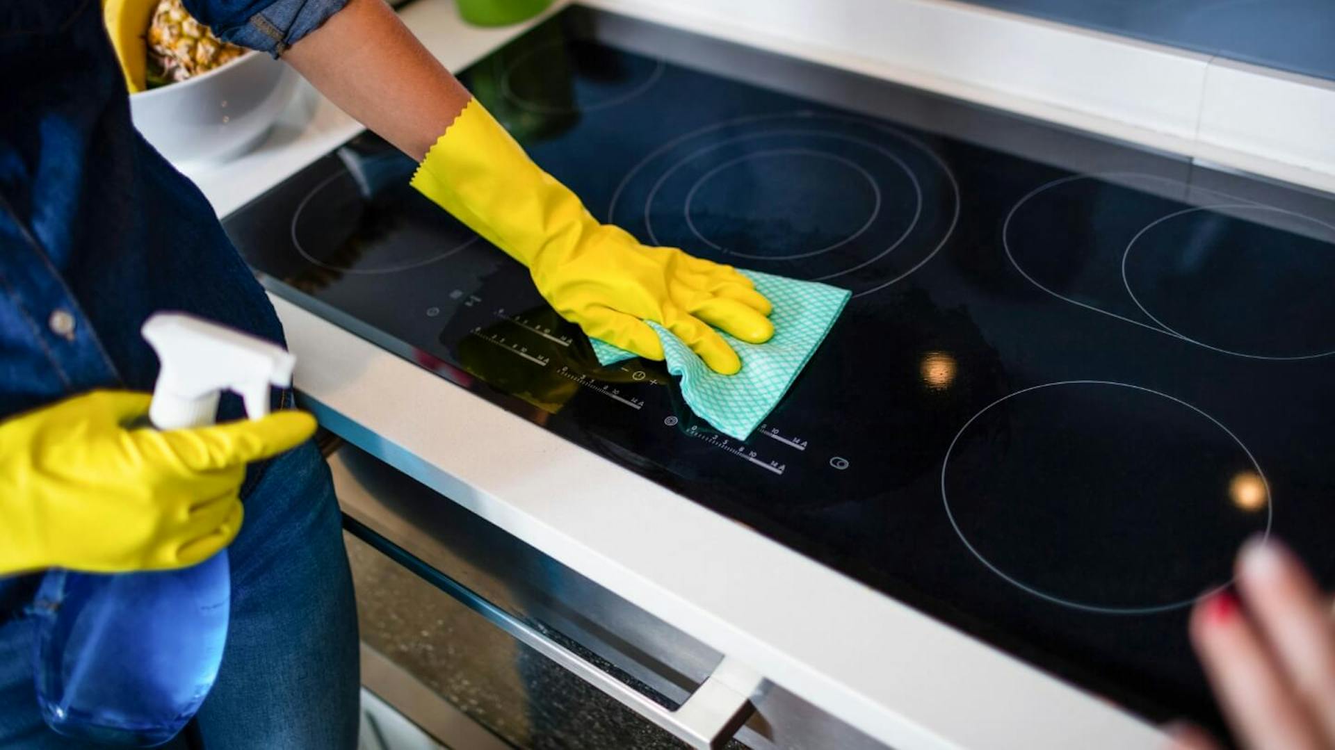 Person mit gelben Gummihandschuhen reinigt mit grünem Lappen Kochfeld.