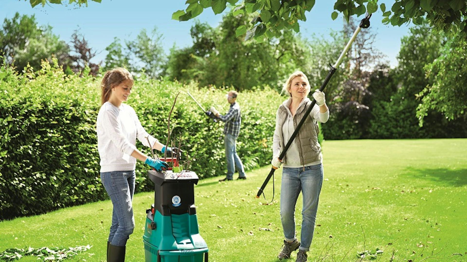 Zwei Frauen im Garten schneiden einen Baum. Die Frau links betätigt einen Häcksler, die Frau recht schneidet den Baum mit einer langen Schere. Im Hintergrund schneidet ein Mann die Hecke.