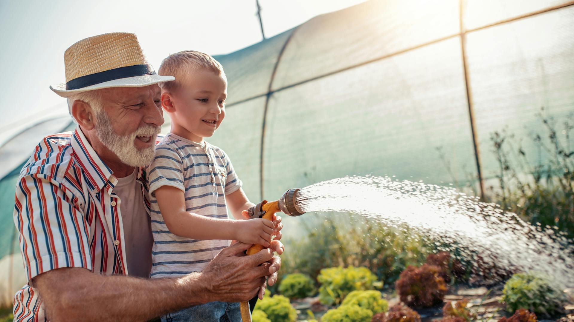 Ein älterer Mann gießt gemeinsam mit seinem Enkel den Garten mit einem Schlauch.